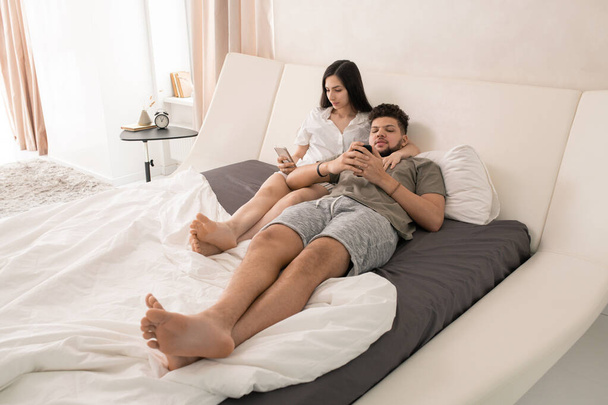 молодая любящая спокойная пара со смартфонами, отдыхающая на большой двуспальной кровати и просматривающая сообщения в социальных сетях - Фото, изображение