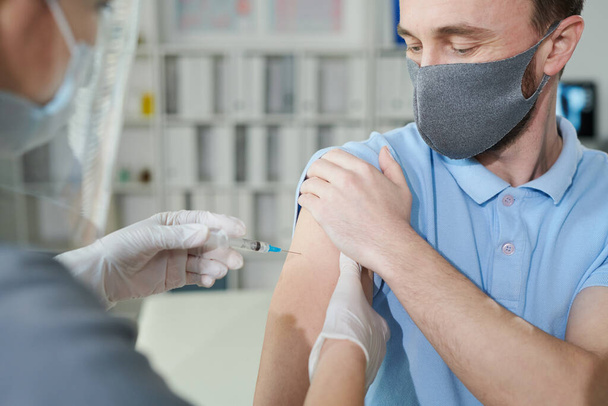 Νεαρός άνδρας κρατά το μανίκι του πουκαμίσου του ενώ ο νοσοκόμος φοράει προστατευτικά ενδύματα εργασίας αναγκάζοντάς τον να κάνει ένεση κατά τη διάρκεια εμβολιασμού κατά του ιού σε κλινικές - Φωτογραφία, εικόνα