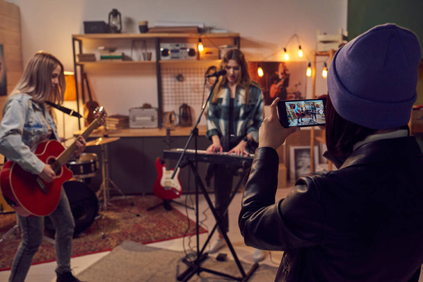 Вид ззаду молодої жінки, що записує виконання своїх друзів на мобільний телефон, граючи на музичних інструментах
 - Фото, зображення