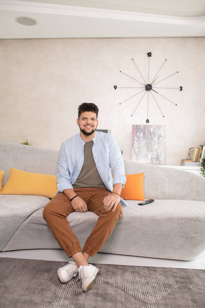 Šťastný mladý vousatý muž v neformálním oblečení se na vás dívá se zubatým úsměvem, zatímco sedí na gauči před kamerou v obývacím pokoji - Fotografie, Obrázek