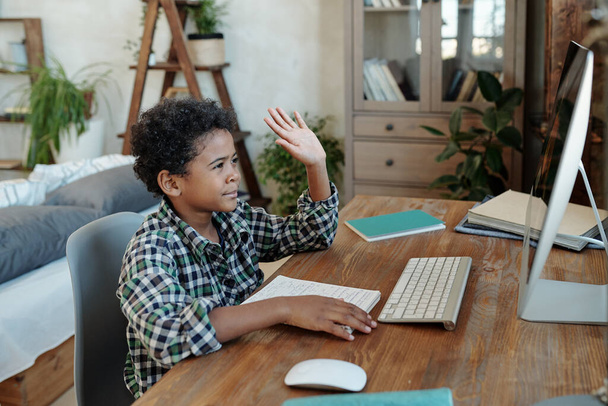glücklicher kleiner Schüler sitzt am Tisch vor dem Computerbildschirm und winkt seinem Lehrer vor Beginn des Online-Unterrichts zu - Foto, Bild