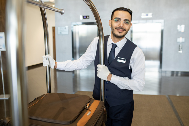 Young porter in uniform and eyeglasses pushing cart with suitcases of guests while moving in front of camera along corridor during work - 写真・画像