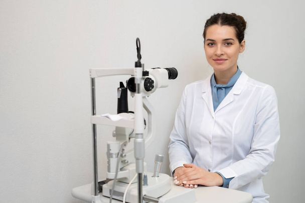 Giovane oftalmologa sorridente in whitecoat seduta davanti alla telecamera da attrezzature mediche in cliniche e in attesa di paziente - Foto, immagini