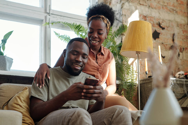 Joyful young African couple in casualwear looking at screen of smartphone held by smiling man while sitting in front of camera against window - 写真・画像