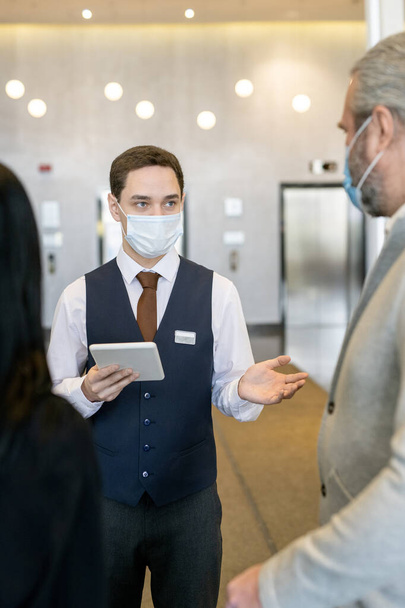 Молодой администратор отеля в защитной маске и формальной одежде с помощью тачпада во время общения с новыми гостями в гостиной - Фото, изображение