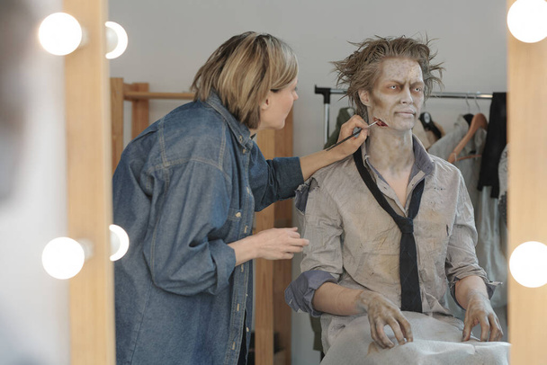 Reflection in mirror of female visage artist with brush applying zombie makeup on face and neck of young businessman or actor in studio - Valokuva, kuva