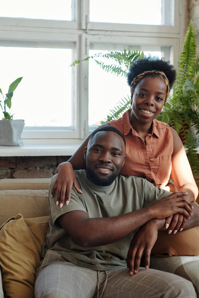 Feliz joven pareja africana en ropa de casualwear mirándote con sonrisas mientras se sienta frente a la cámara contra la ventana con plantas domésticas verdes - Foto, Imagen