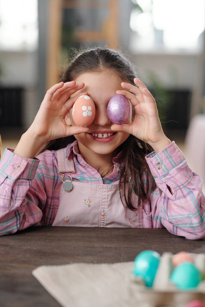 Menina bonito alegre em casualwear segurando pintado ovos de Páscoa por seus olhos enquanto sentado por grande mesa de madeira em ambiente doméstico - Foto, Imagem
