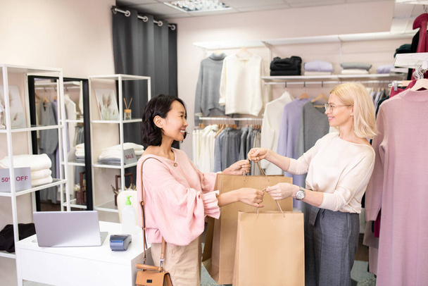 Asiatische junge Kundin kauft Kleidung im Laden, während Verkäuferin lächelnd ihre Einkaufstüten reicht - Foto, Bild