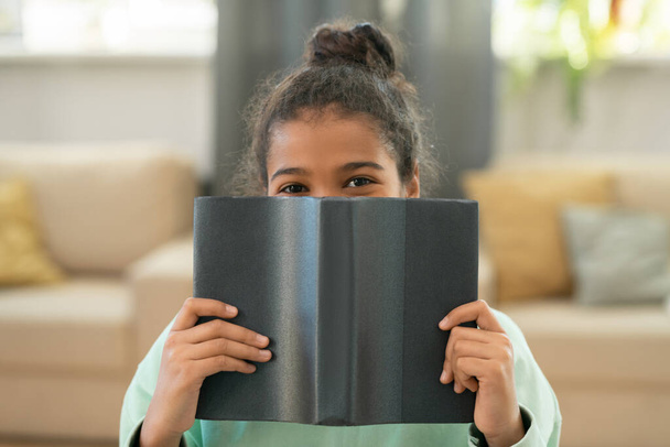 Cute elementary schoolgirl sitting in front of camera in living-room against couch with pillows and peeking at you out of open book - Fotoğraf, Görsel