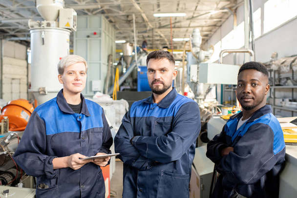 Drie jonge serieuze arbeiders van de hedendaagse fabriek in overalls voor de camera tegen industriële machines in een grote werkplaats - Foto, afbeelding
