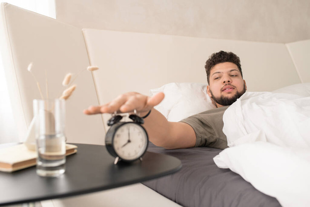 молодой бородатый мужчина в футболке, нажимая кнопку будильника, лежа под белым одеялом в большой кровати после сна утром - Фото, изображение