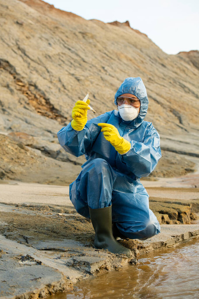 Female ecologist in gloves, coveralls, respirator and eyeglasses looking at flask with sample of dirty water or soil in dangerously polluted area - Fotó, kép