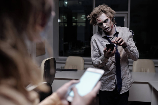 Homem de negócios morto e assustador com tinta gordurosa zumbi no rosto e mãos rolando no smartphone no escritório à meia-noite com a fêmea na frente - Foto, Imagem