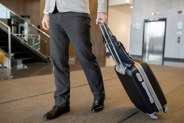 Low section of modern businessman in casualwear pulling suitcase behind himself while moving along hall of large and luxurious hotel - Photo, Image