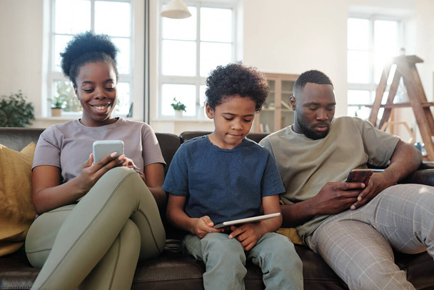 Familia joven de padre, madre e hijo pequeño lindo usando gadgets móviles mientras está sentado en fila en el sofá de cuero negro en el entorno del hogar - Foto, imagen
