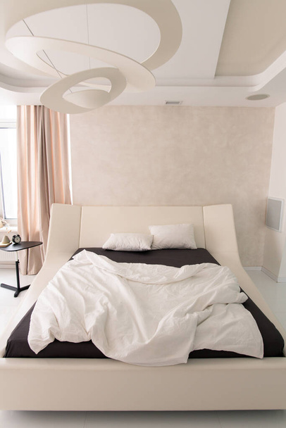 Interieur van gezellige slaapkamer met comfortabel tweepersoonsbed met geometrische kroonluchter boven, wekker op tafeltje en beige gordijn op raam - Foto, afbeelding