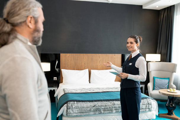 Young smiling hotel worker in uniform holding digital tablet while showing mature businessman his room and wishing him good rest - Fotografie, Obrázek