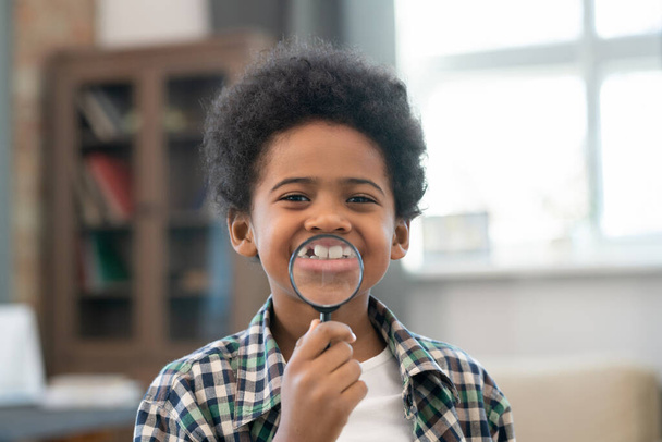 Adorable schoolboy of African ethnicity holding magnifying glass in front of his mouth and showing his toothy smile while looking at you - Foto, immagini