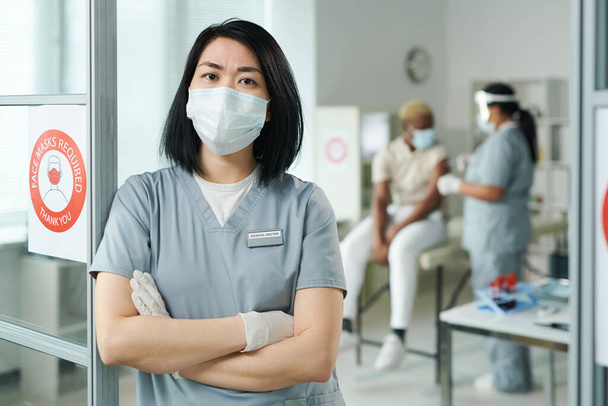 junge, kreuzarmige Ärztin in Schutzkleidung steht vor der Kamera, während ihr Kollege eine Patientin impft - Foto, Bild