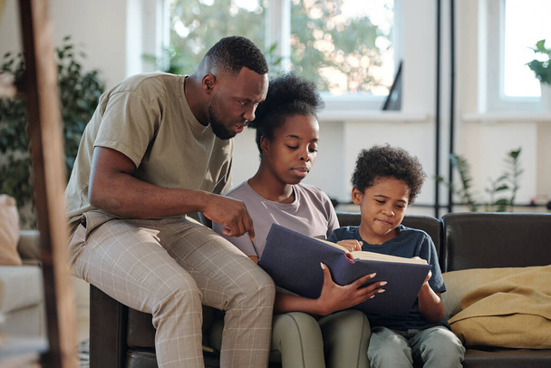Jovem família africana de pais e pequeno filho bonito sentado no sofá na sala de estar e lendo grande livro de contos ou enciclopédia - Foto, Imagem