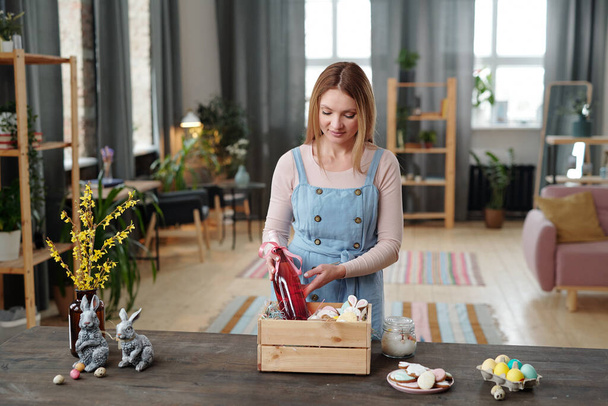 Young blond female putting bottle of fruit drink for children and other snacks into wooden box while standing by table in living-room - Foto, imagen