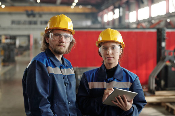 Dos jóvenes ingenieros serios en sombreros protectores y ropa de trabajo que te miran mientras estás parado frente a la cámara dentro de un gran taller - Foto, Imagen
