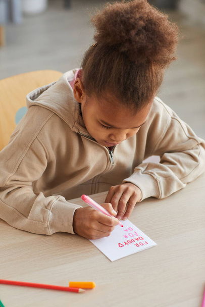 Retrato vertical de una linda chica afroamericana haciendo una tarjeta hecha a mano como regalo para el día de los padres, espacio para copiar - Foto, imagen