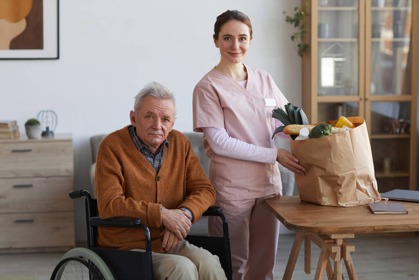 Ritratto di uomo anziano in sedia a rotelle in posa con giovane badante donna in possesso di sacchetto di generi alimentari, sia guardando la fotocamera, spazio copia - Foto, immagini
