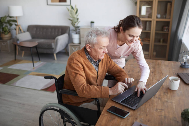 Retrato del hombre mayor en silla de ruedas usando el portátil en el hogar de retiro con la enfermera que lo asiste, espacio de copia - Foto, imagen
