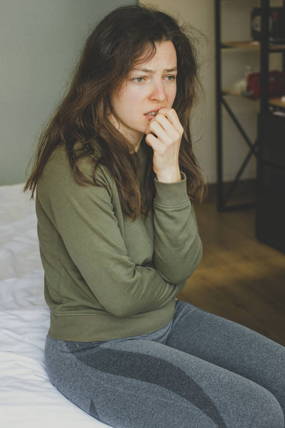 Worried sad young brunette woman thinking about problems, biting nails sitting on bed at home interior. Unhappy teenager suffering from psychological problem, unexpected pregnancy concept. Vertical. - Photo, Image