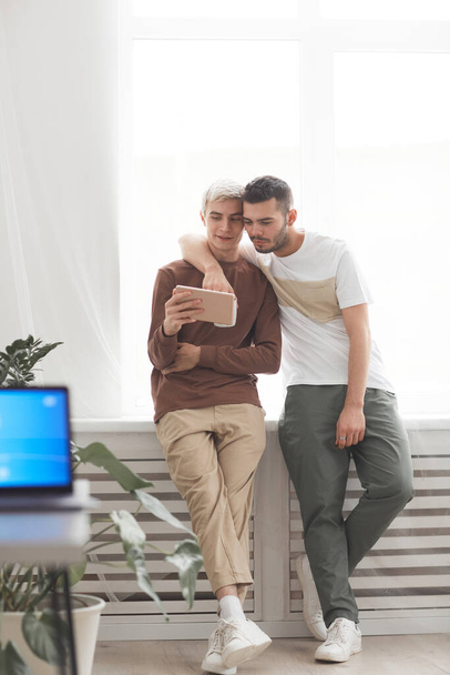 Вертикальный портрет современной пары геев, обнимающихся дома, стоя у окна и используя цифровые планшеты - Фото, изображение