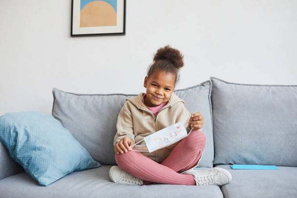 Full length portrait of cute African-American girl sitting cross-legged on sofa and smiling at camera on Fathers day, copy space - Zdjęcie, obraz