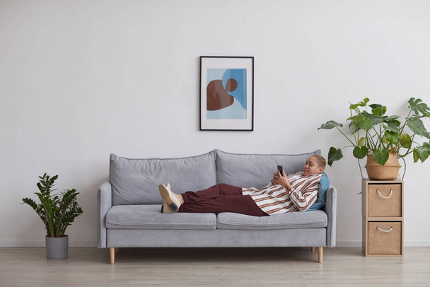 Wide angle view at modern mixed-race woman using smartphone while lying on sofa in minimal home interior, copy space - Foto, Imagen