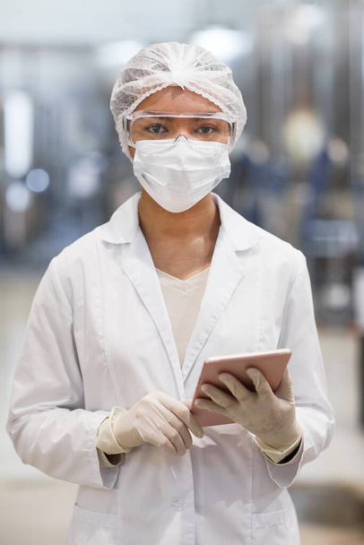 Κάθετη μέση προς τα πάνω πορτρέτο της νεαρής γυναίκας που εργάζεται σε χημικό εργοστάσιο και κοιτάζοντας την κάμερα φορώντας προστατευτικό ρουχισμό - Φωτογραφία, εικόνα