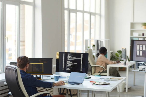 Achtergrond beeld van IT-ontwikkelaars kantoor met focus op mannelijke programmeur schrijven code op computerscherm, kopieer ruimte - Foto, afbeelding