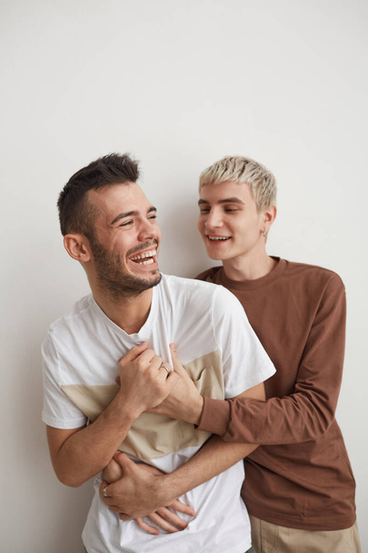 verticaal portret van zorgeloos gay paar omarmen en lachen gelukkig terwijl poseren tegen wit in huis - Foto, afbeelding