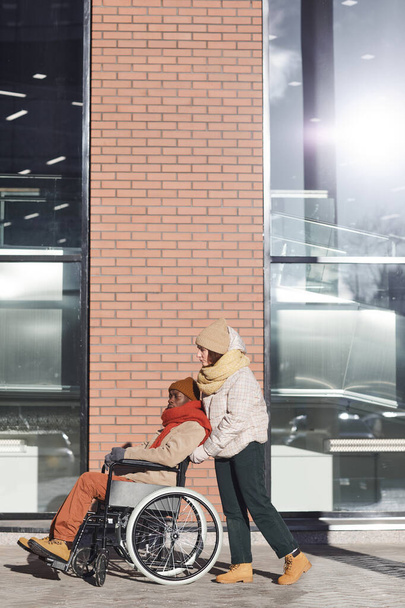 Vertikales Ganzkörperporträt eines afroamerikanischen Mannes im Rollstuhl, der sich in einer städtischen Stadt bewegt und dabei von einer jungen Frau unterstützt wird, Kopierraum - Foto, Bild