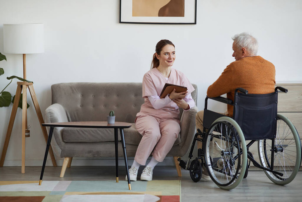 Retrato completo de enfermeira sorridente falando com homem idoso em cadeira de rodas e usando tablet digital em casa de repouso, espaço de cópia - Foto, Imagem