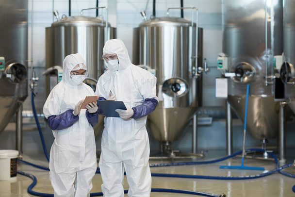 Modern kimya fabrikasında dijital tablet kullanırken tehlikeli madde giysileri giyen iki genç işçinin belden yukarı portresi. - Fotoğraf, Görsel