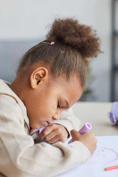Vue latérale verticale portrait de fille afro-américaine mignonne dessinant des images tout en étant assis à la table à la maison, espace de copie - Photo, image