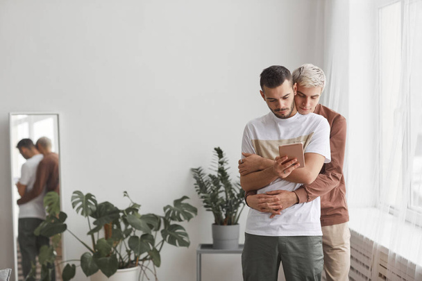 Retrato de vista frontal de pareja gay contemporánea abrazando mientras mira la pantalla de la tableta en el interior del hogar mínimo, espacio de copia - Foto, imagen