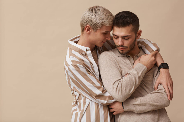 Minimal μέση προς τα πάνω πορτρέτο της αγάπης γκέι ζευγάρι αγκαλιάζει ενώ θέτουν σε ουδέτερο μπεζ φόντο στο στούντιο, αντιγραφή χώρου - Φωτογραφία, εικόνα