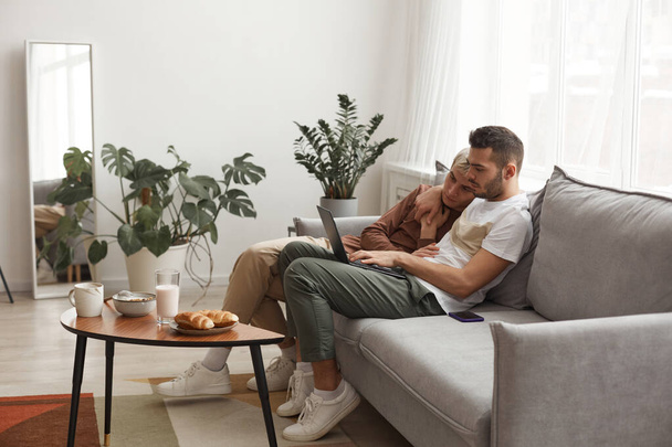 Вид сбоку на современную гей-пару в повседневной жизни, двое молодых людей, использующих ноутбук вместе, расслабляясь на диване дома, пространство для копирования - Фото, изображение