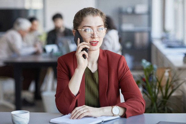 Portrait of young elegant businesswoman speaking by phone while working at desk in office with team in background - Фото, зображення