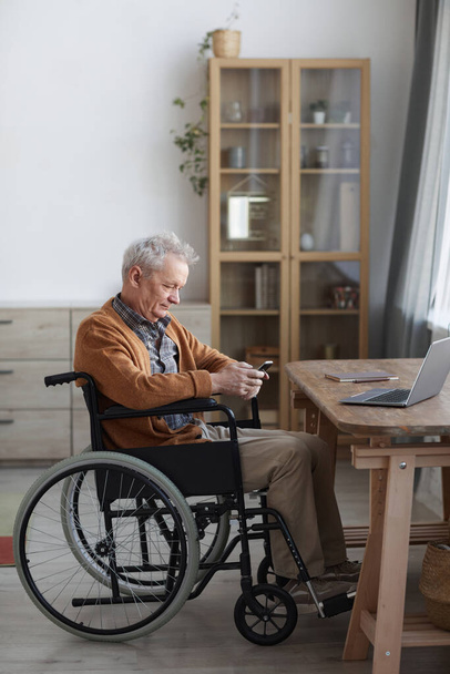 Retrato vertical de vista lateral del hombre mayor en silla de ruedas usando un teléfono inteligente mientras está sentado junto al escritorio en el interior del hogar - Foto, imagen