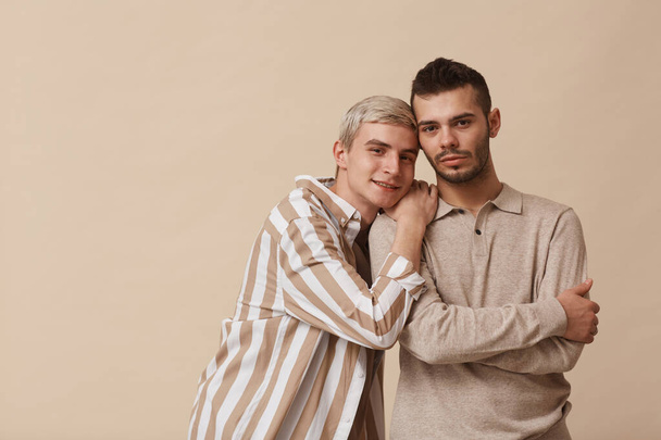 Cintura mínima até retrato de feliz casal gay olhando para a câmera enquanto posando contra fundo bege em estúdio, espaço de cópia - Foto, Imagem