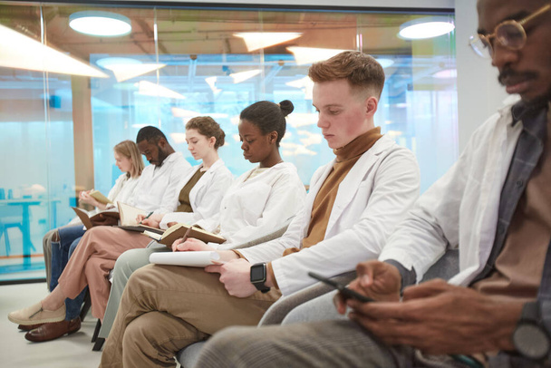 Multi-ethnic group of people wearing lab coats while sitting in row and taking notes during lecture on medicine in college, copy space - Foto, Imagen