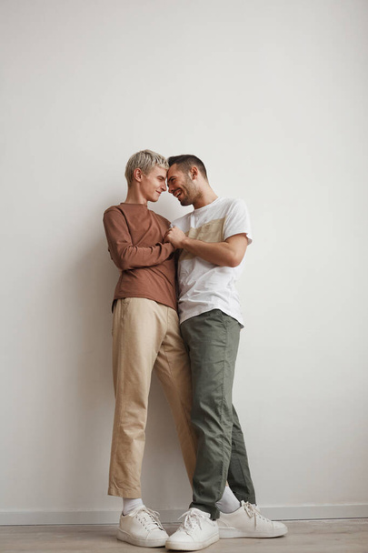 Вертикальный полноразмерный портрет влюбленной гей-пары, обнимающейся дома, стоя у белой стены, копирующее пространство - Фото, изображение