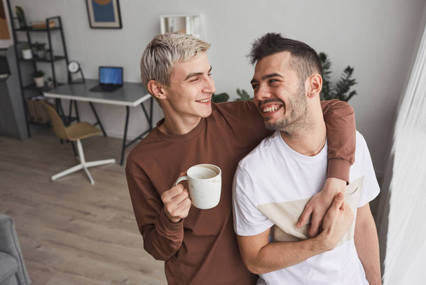 Candid portrait of carefree gay couple embracing while standing by window and enjoying morning together at home, copy space - Foto, Imagen